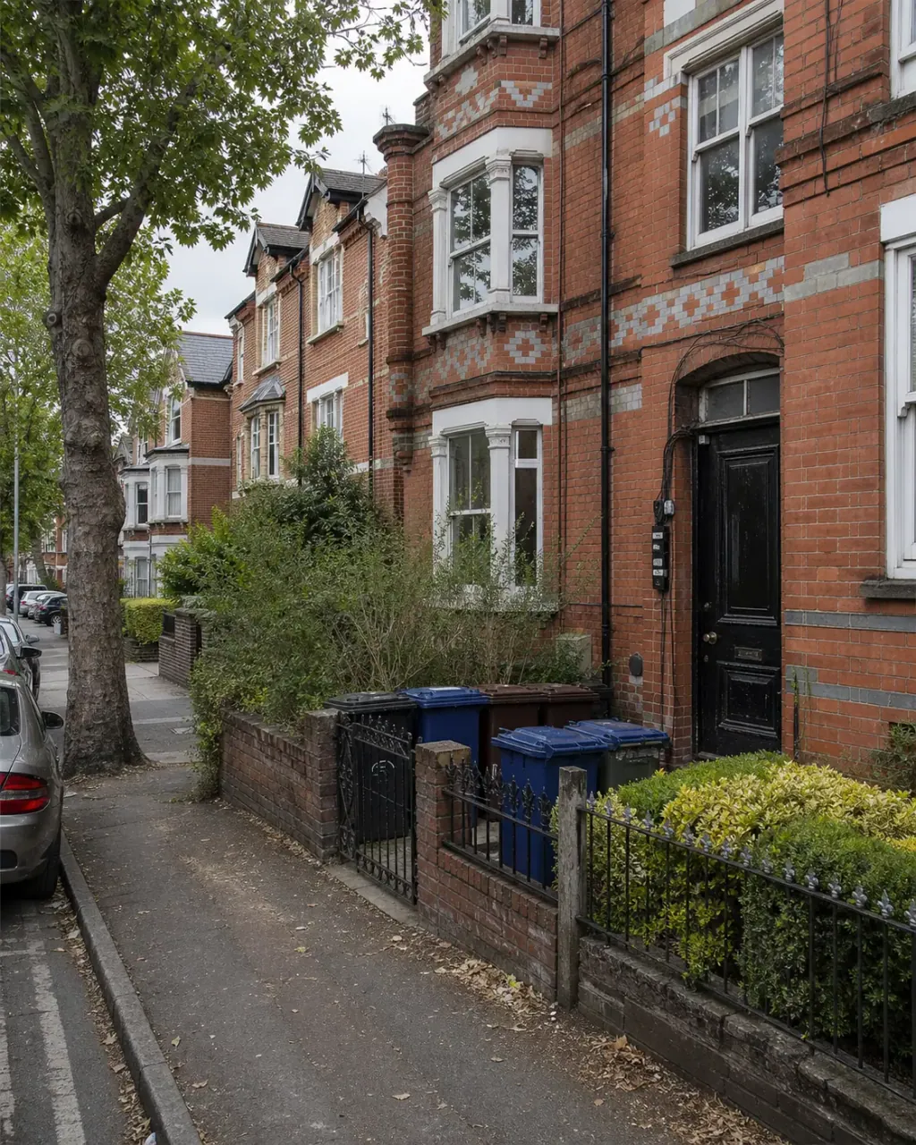 Three-storey red-brick Victorian terraces with grey chequer brickwork on a tree-lined Redlands street, recycling bins grouped by a front gate
