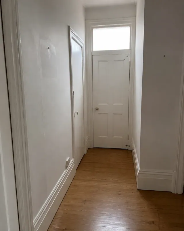 The bare hallway of a conversion flat looking to a white panelled front door, floorboards swept, one picture hook left on the wall