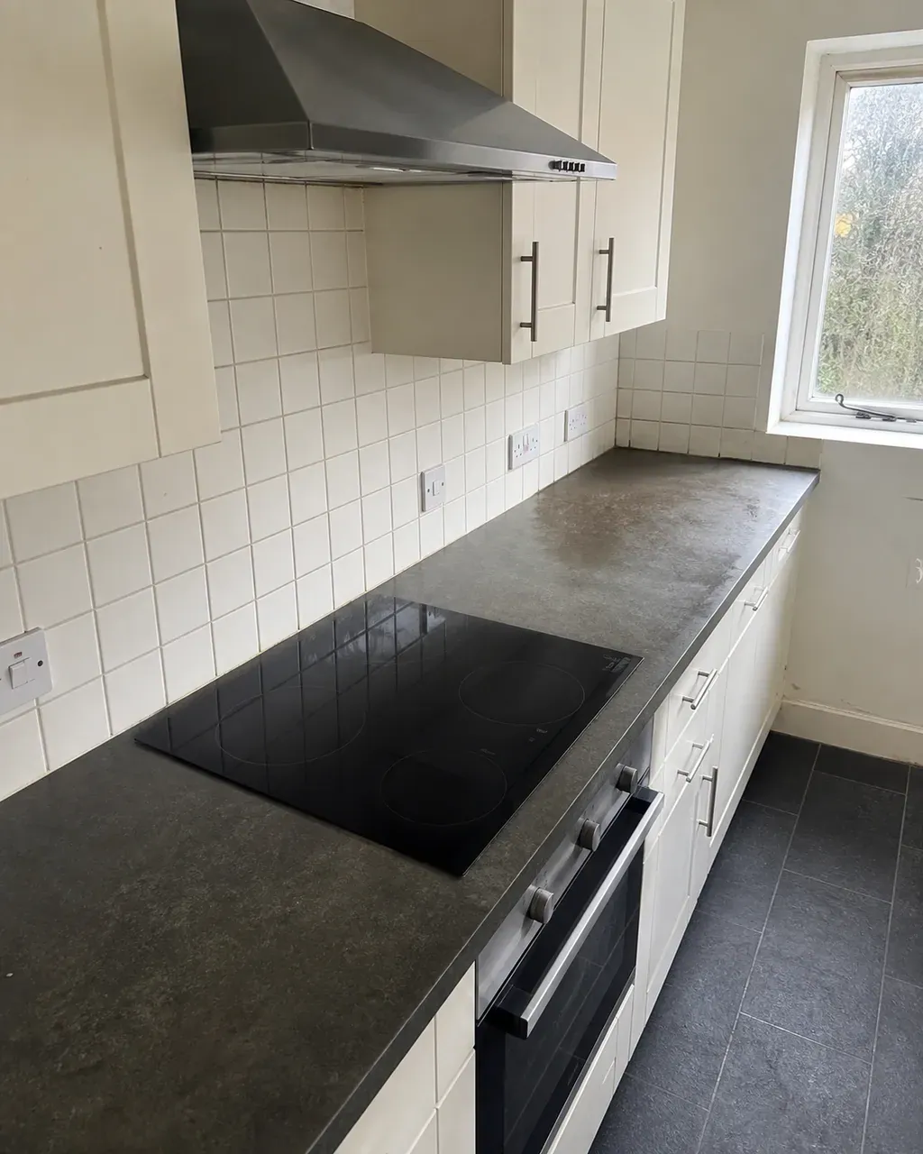 A galley kitchen worktop and induction hob polished for checkout, white tiles and extractor hood catching the window light